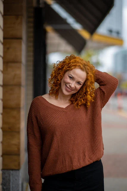 Lächelnde rothaarige Person mit Locken und braunem Pullover, die mit einer Hand auf dem Kopf im Freien steht; städtischer Hintergrund.