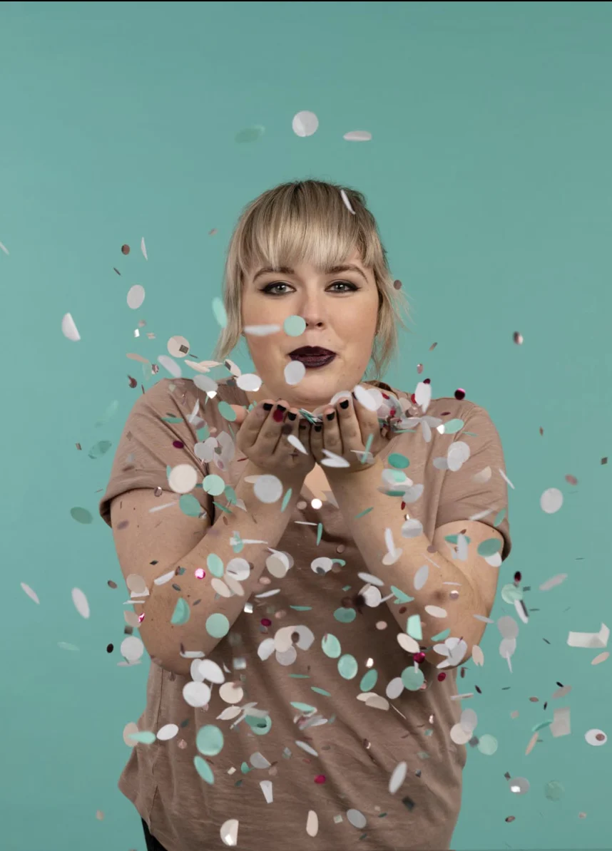 Blonde Frau in taupem Shirt bläst weißen, teal- und metallicfarbenen Konfetti auf Kamera vor teal Hintergrund. Dunkler Lippenstift.