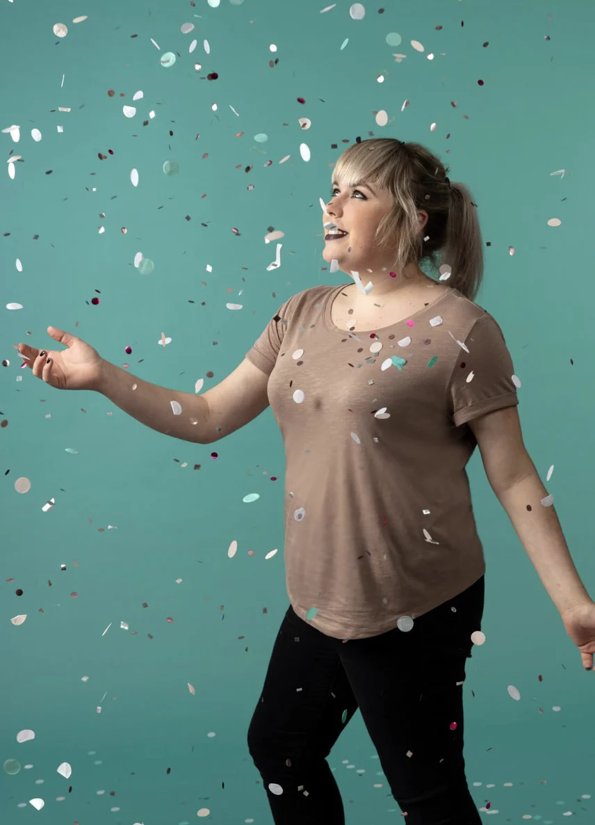 Person mit blonden Haaren und braunem T-Shirt lächelt vor blaugrünem Hintergrund, während bunte Konfetti fallen.
