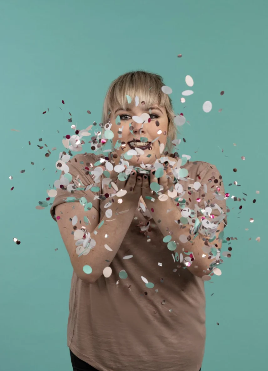 Person mit blondem Haar in braunem Shirt bläst buntes Konfetti gegen die Kamera vor einem türkisfarbenen Hintergrund.