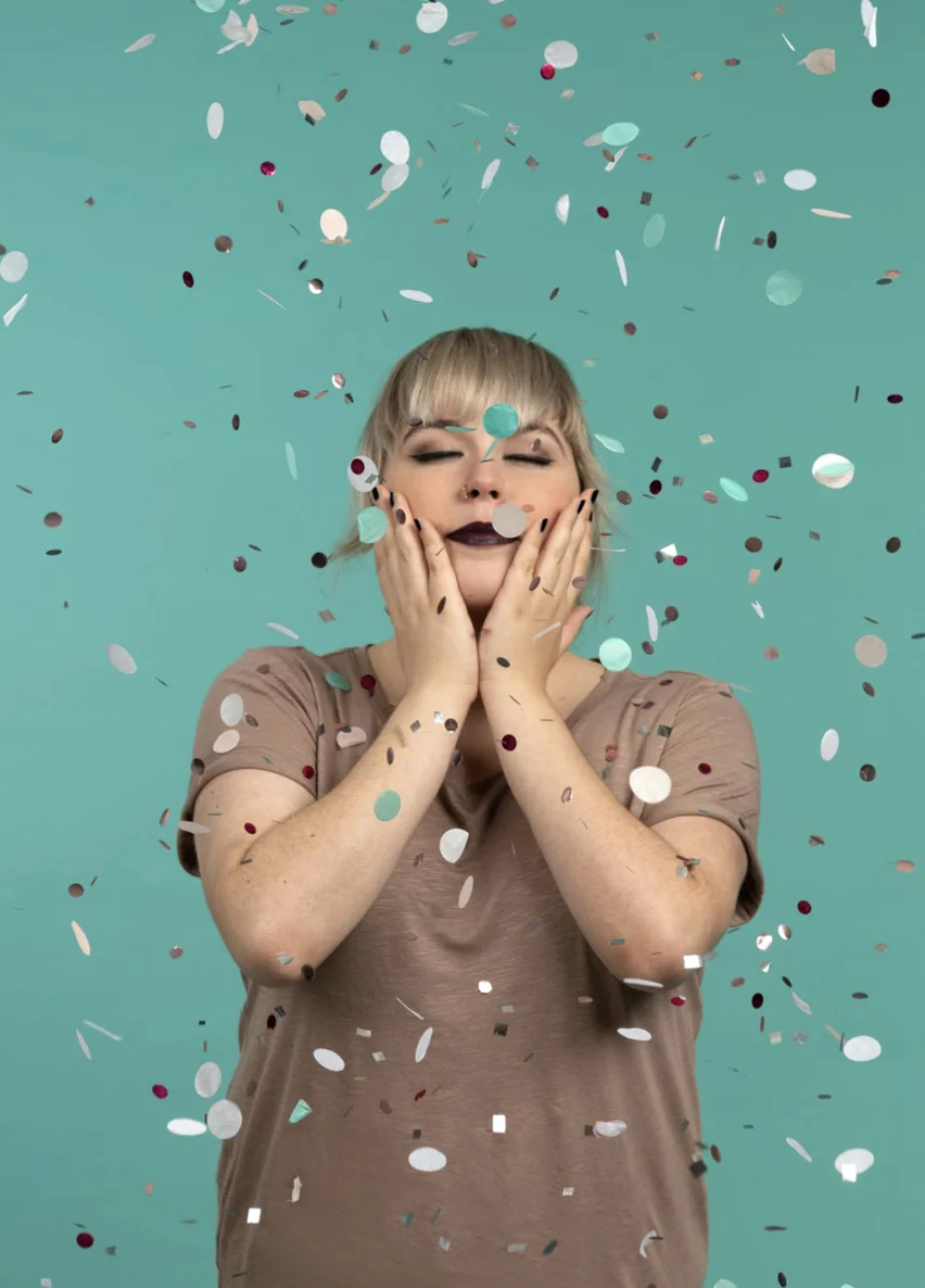 Person mit entspanntem Gesicht berührt sanft ihr Gesicht; blaugrüner Hintergrund, fallendes Konfetti, beiges Hemd.
