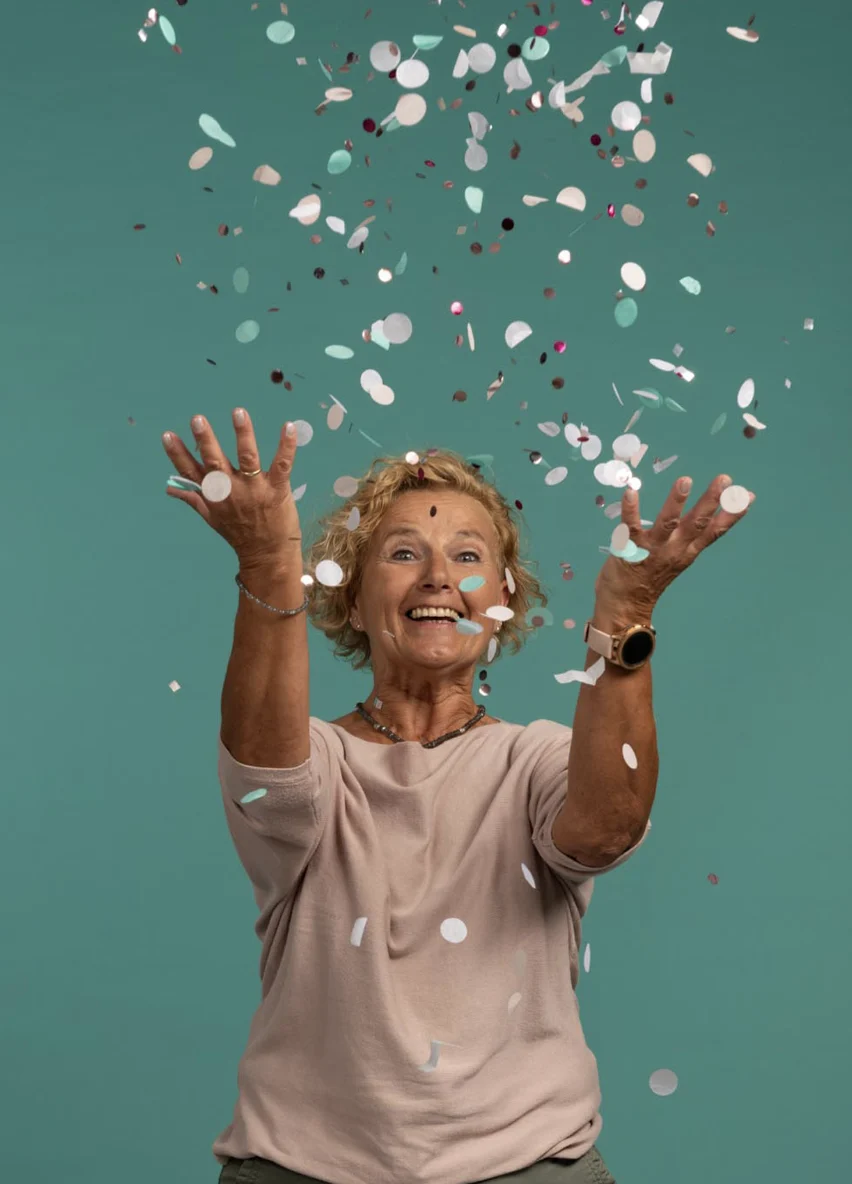 Eine fröhliche Frau mit kurzen lockigen Haaren wirft buntes Konfetti, lächelt vor einem blaugrünen Hintergrund und trägt ein rosa Oberteil.
