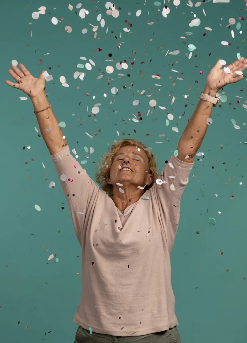 Eine Person mit Locken und rosa Hemd wirft freudig Konfetti vor einem blaugrünen Hintergrund, hat die Arme erhoben und blickt zwischen herabfallendem Konfetti nach oben.