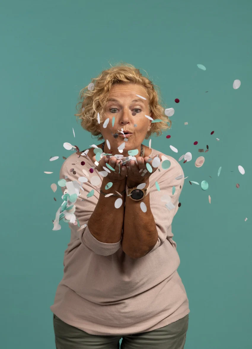 Ältere Frau mit lockigem Haar bläst spielerisch buntes Konfetti, trägt rosa Pullover, vor einem tealfarbenen Hintergrund.