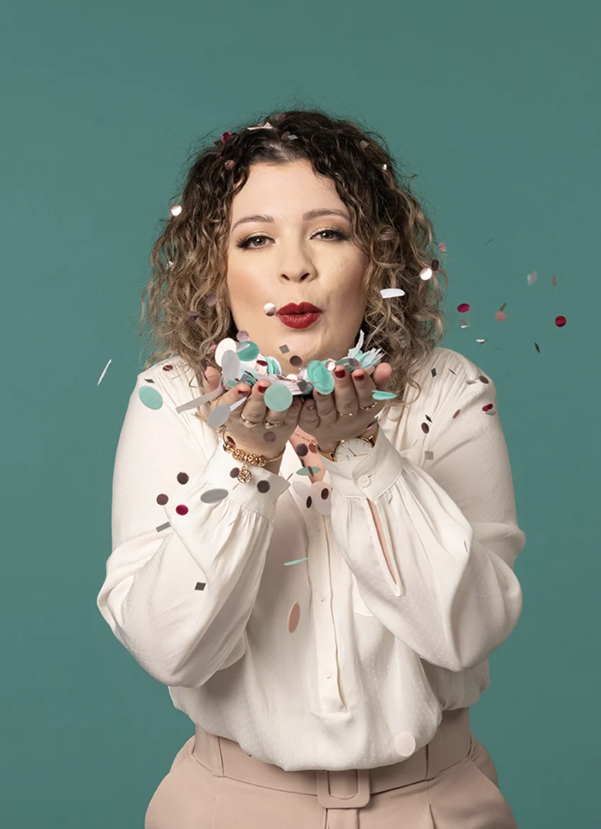 Frau mit lockigem Haar bläst Konfetti vor einem türkisfarbenen Hintergrund, trägt weiße Bluse, beige Hose und wirkt fröhlich.