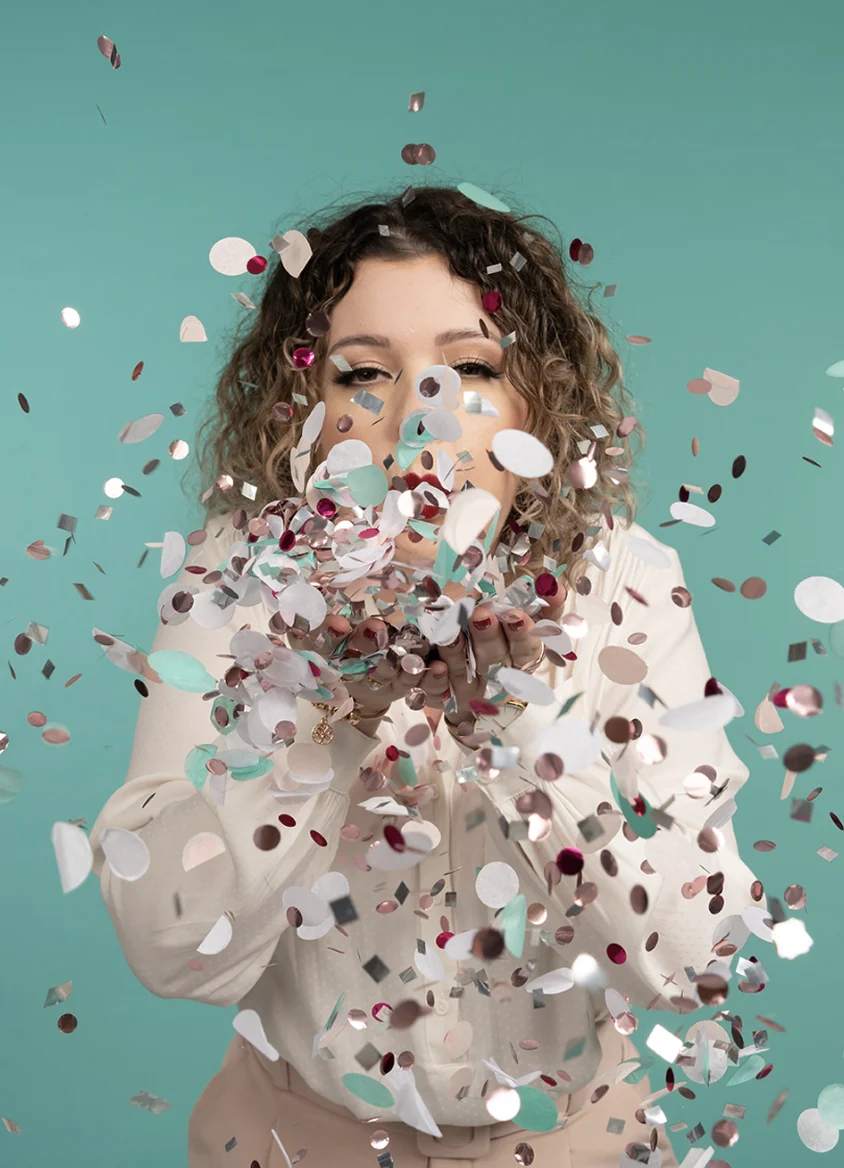 Person mit lockigem Haar bläst buntes Konfetti zur Kamera vor einem türkisfarbenen Hintergrund, trägt ein weißes Hemd. Feierstimmung.