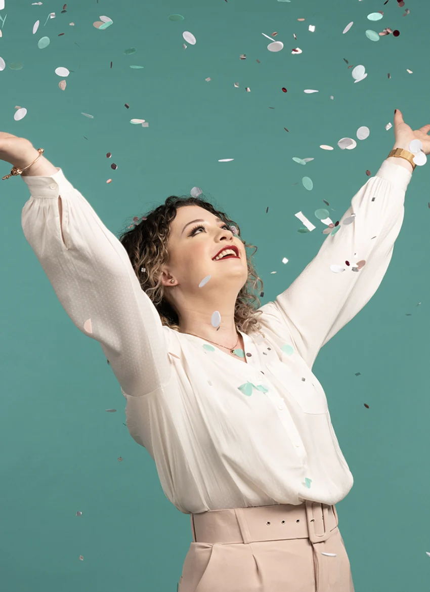Frau mit lockigem Haar, weißer Bluse und beiger Hose wirft fröhlich buntes Konfetti vor einem blaugrünen Hintergrund.