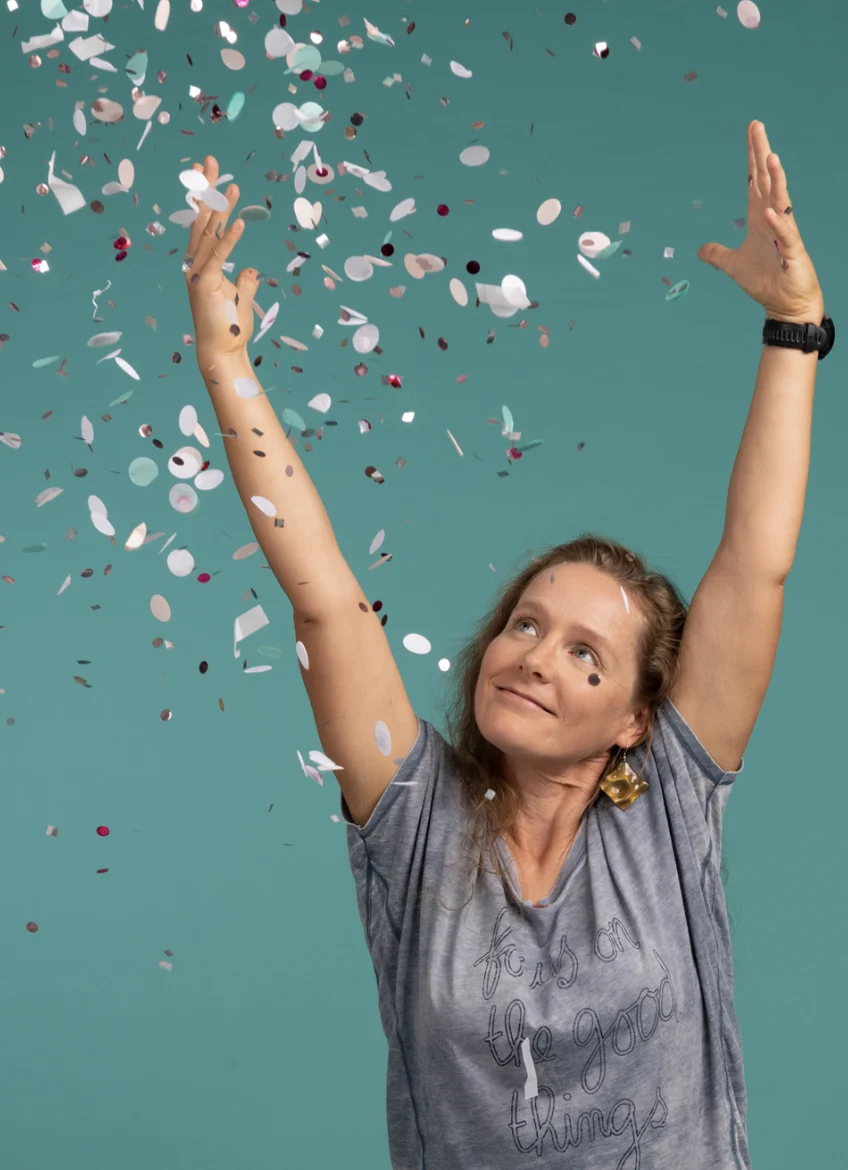 Eine fröhliche Frau in einem grauen T-Shirt und Ohrringen wirft Konfetti vor einem blaugrünen Hintergrund.