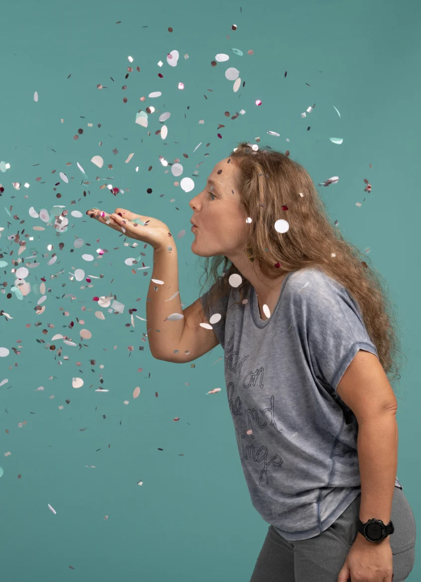 Frau mit langen Haaren bläst vor einem blaugrünen Hintergrund buntes Konfetti aus ihrer Hand.
