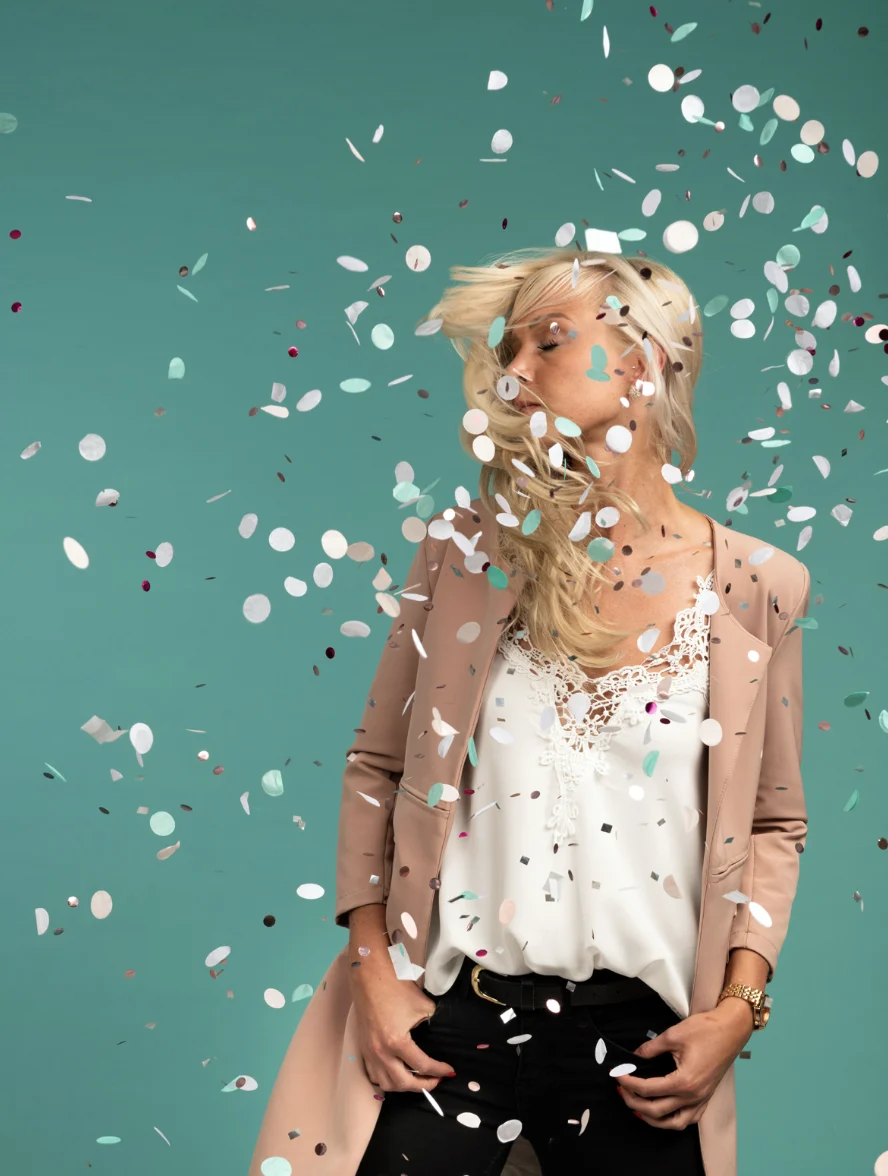 Person mit langen blonden Haaren, beigem Blazer, weißem Spitzenoberteil, Händen in schwarzen Hosentaschen, buntem Konfetti vor türkisfarbenem Hintergrund.