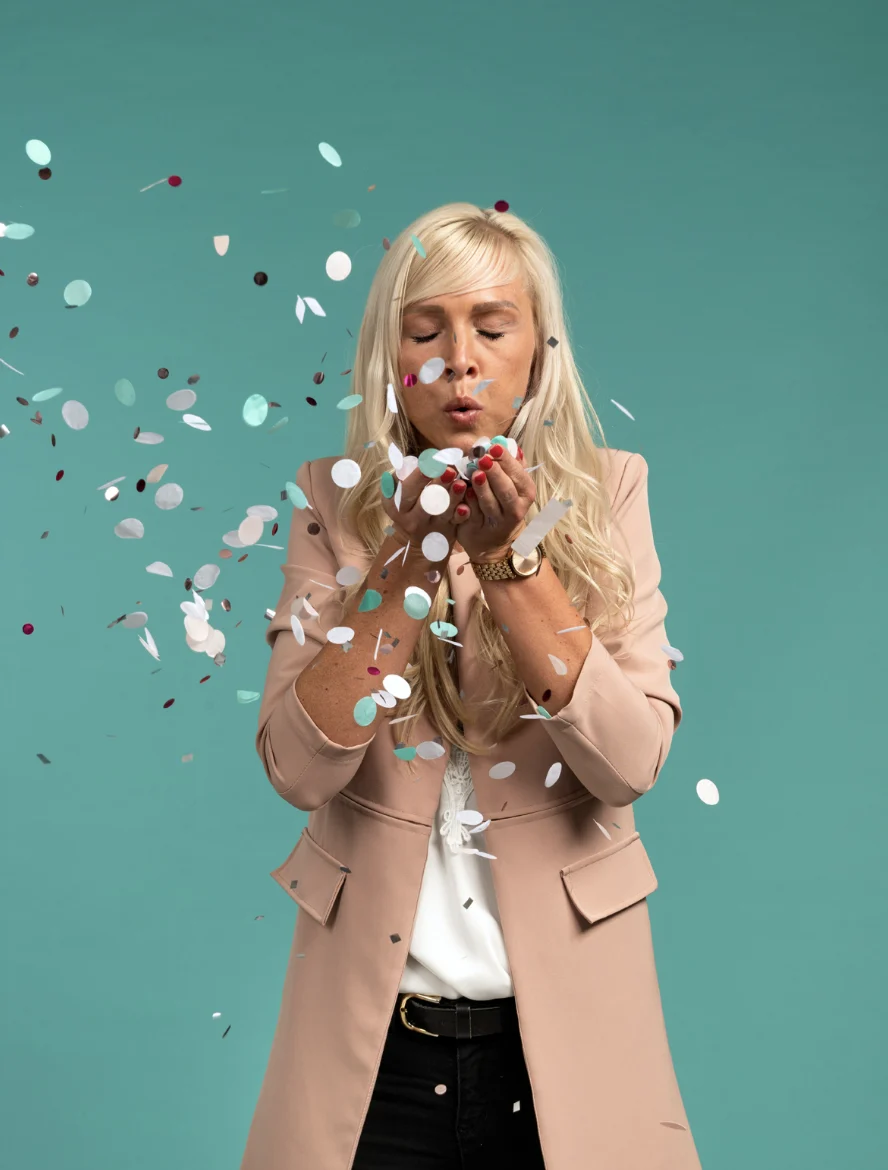 Eine fröhliche blonde Frau in einem beigen Blazer bläst vor einem blaugrünen Hintergrund buntes Konfetti in Richtung Kamera.