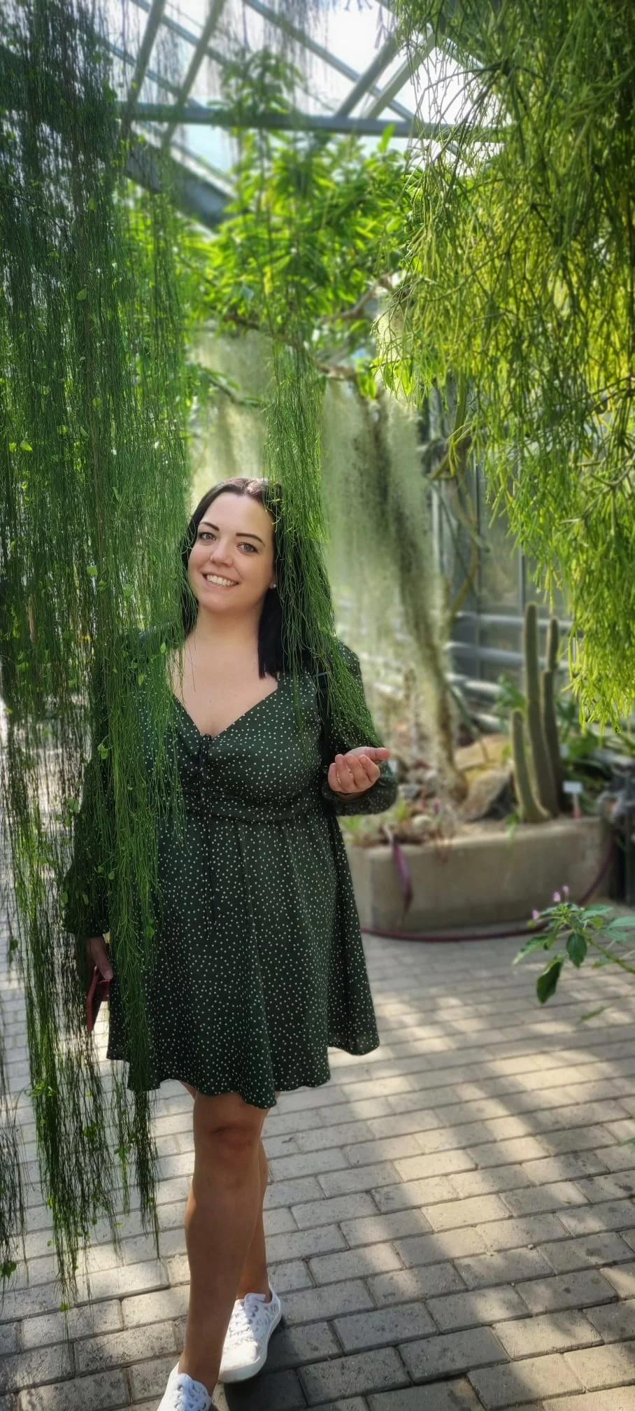 Eine lächelnde Frau in einem grünen Kleid hält ein Blatt in einem Gewächshaus, umgeben von Pflanzen und Kakteen, und trägt weiße Turnschuhe.