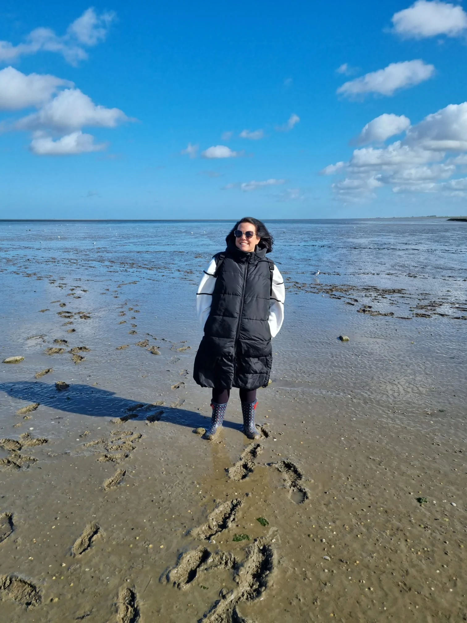 Person im langen schwarzen Mantel und Sonnenbrille am sandigen Strand mit flachem Wasser, blauem Himmel und Fußspuren.