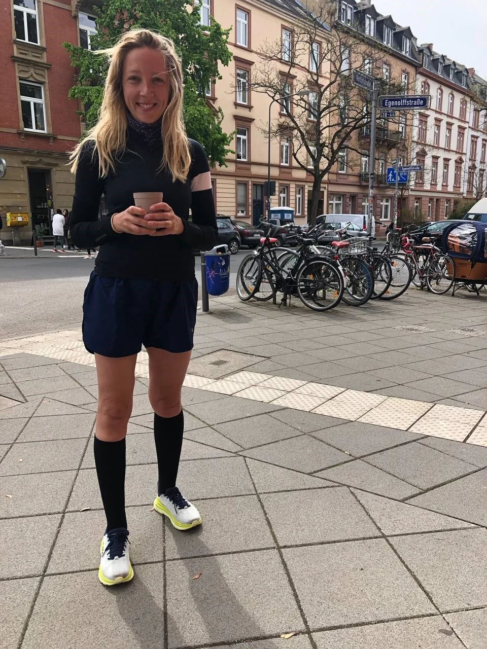 Frau in Sportkleidung hält Tasse auf Stadtgehsteig; schwarzes Oberteil, blaue Shorts, Kniestrümpfe, weiße Turnschuhe; Fahrräder/Gebäude im Hintergrund.