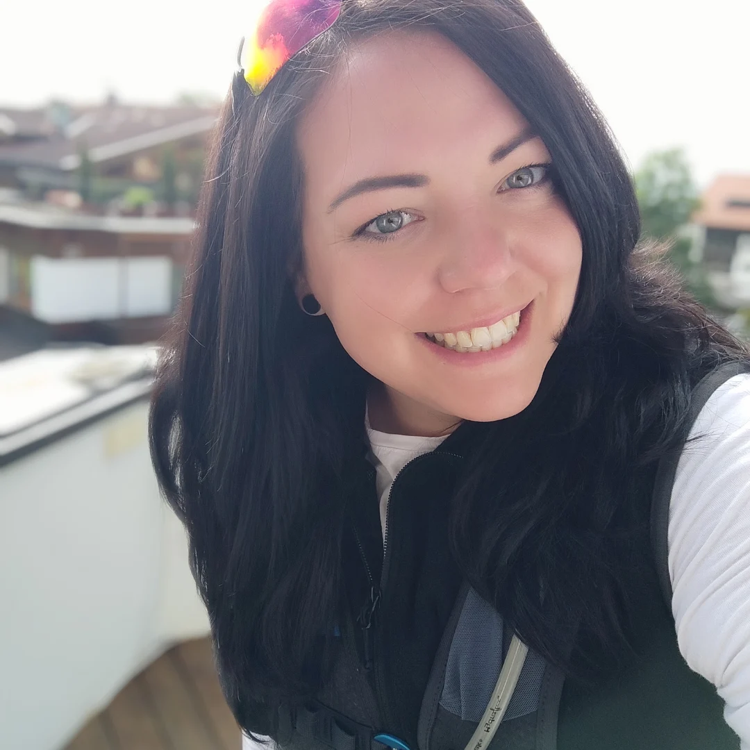 Eine lächelnde Frau mit langen dunklen Haaren und Sonnenbrille auf dem Kopf, in einer schwarzen Weste. Im Hintergrund sind Gebäude verschwommen.