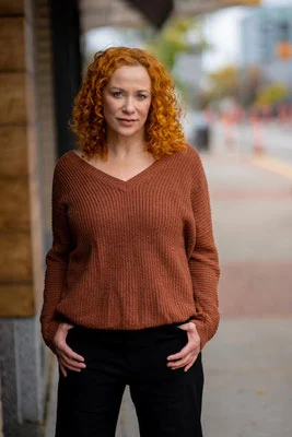 Rothaarige Person in rostrotem Pullover und schwarzer Hose steht mit den Händen in den Taschen auf einem Bürgersteig; städtischer Hintergrund.