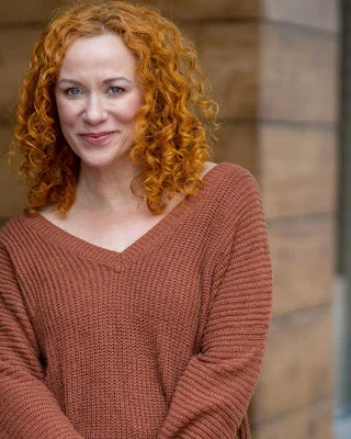 Frau mit lockigem roten Haar, braunem Pullover, steht lächelnd vor Holzwand.