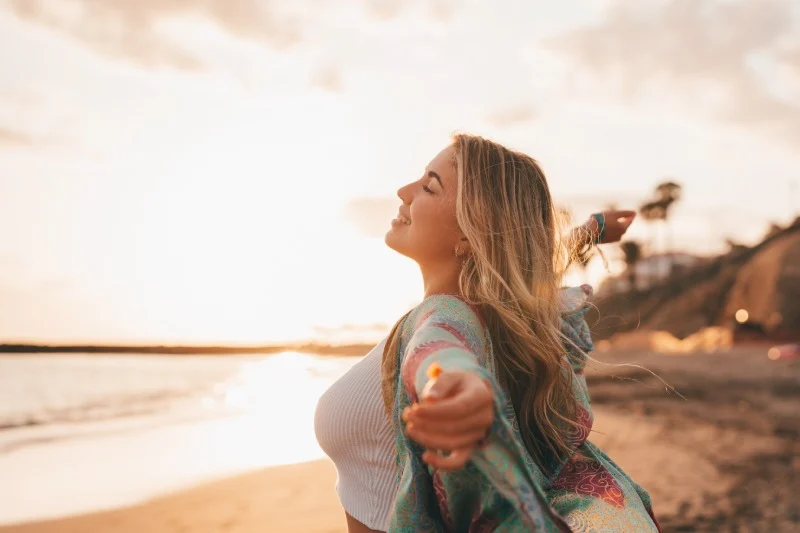 Frau mit langem Haar und farbigem Schal steht am Strand bei Sonnenuntergang, Arme ausgebreitet, reflektiert über Lipödem-Erfahrungen.