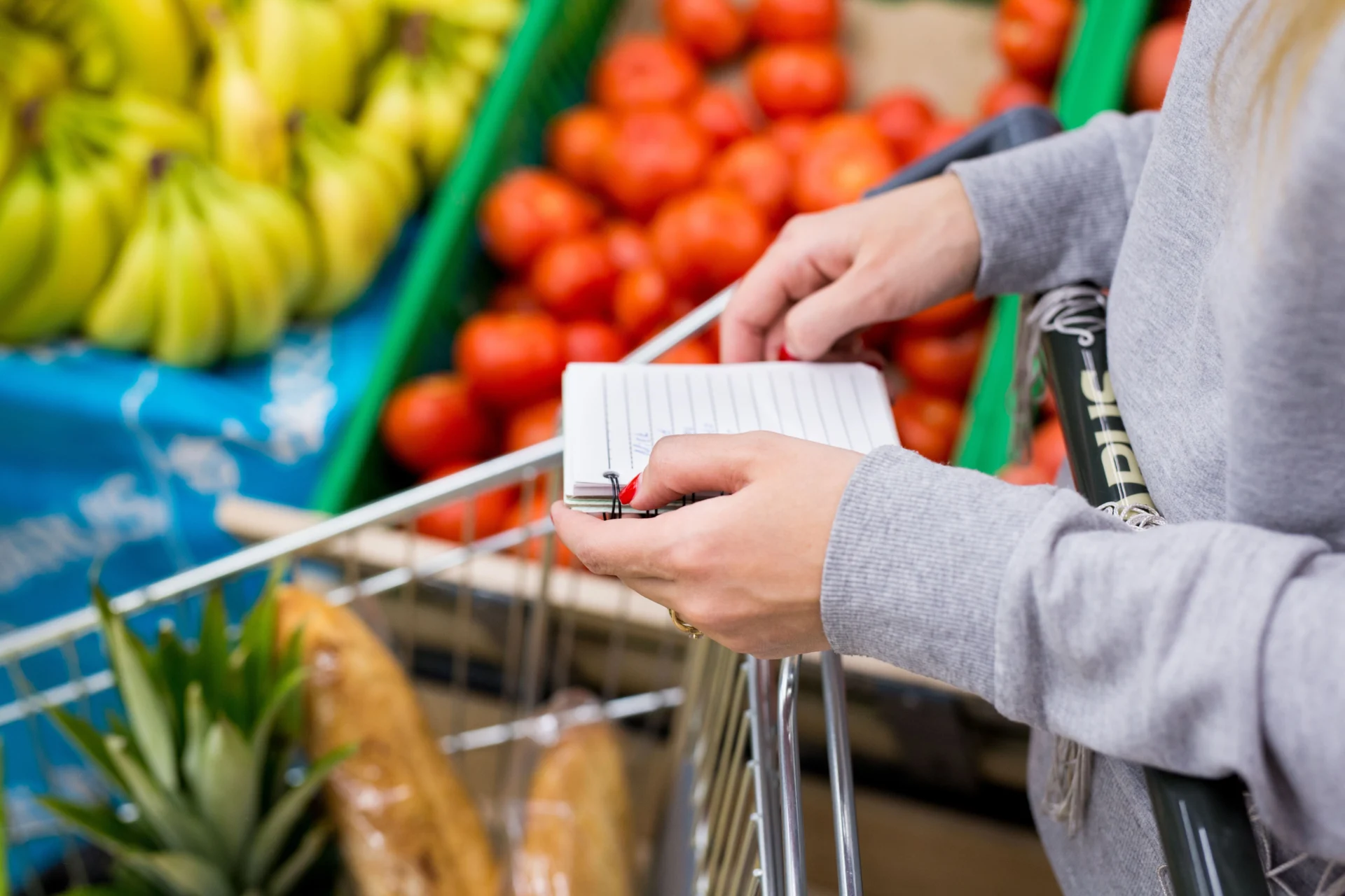 Person neben Einkaufswagen mit Ananas und Baguettes, hält Einkaufsliste und Stift; grauer Pullover, konzentriert.