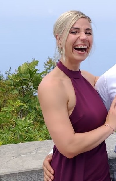 Blonde Frau mit Hochsteckfrisur, lacht in lila Kleid vor grüner Kulisse und klarem Himmel.