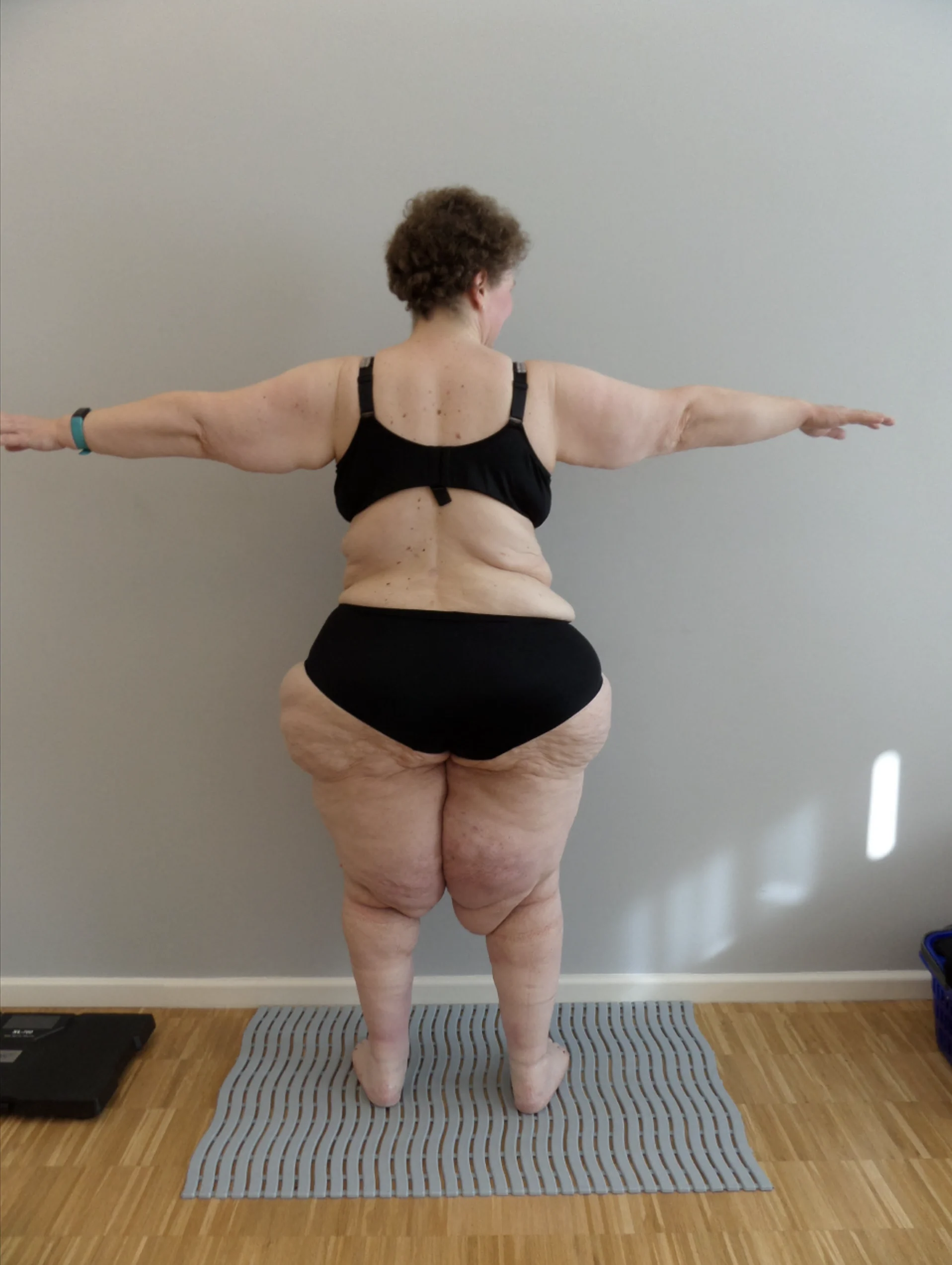 Person mit ausgestreckten Armen, in schwarzem Sport-BH und Shorts, auf heller Matte vor grauem Wand stehend.