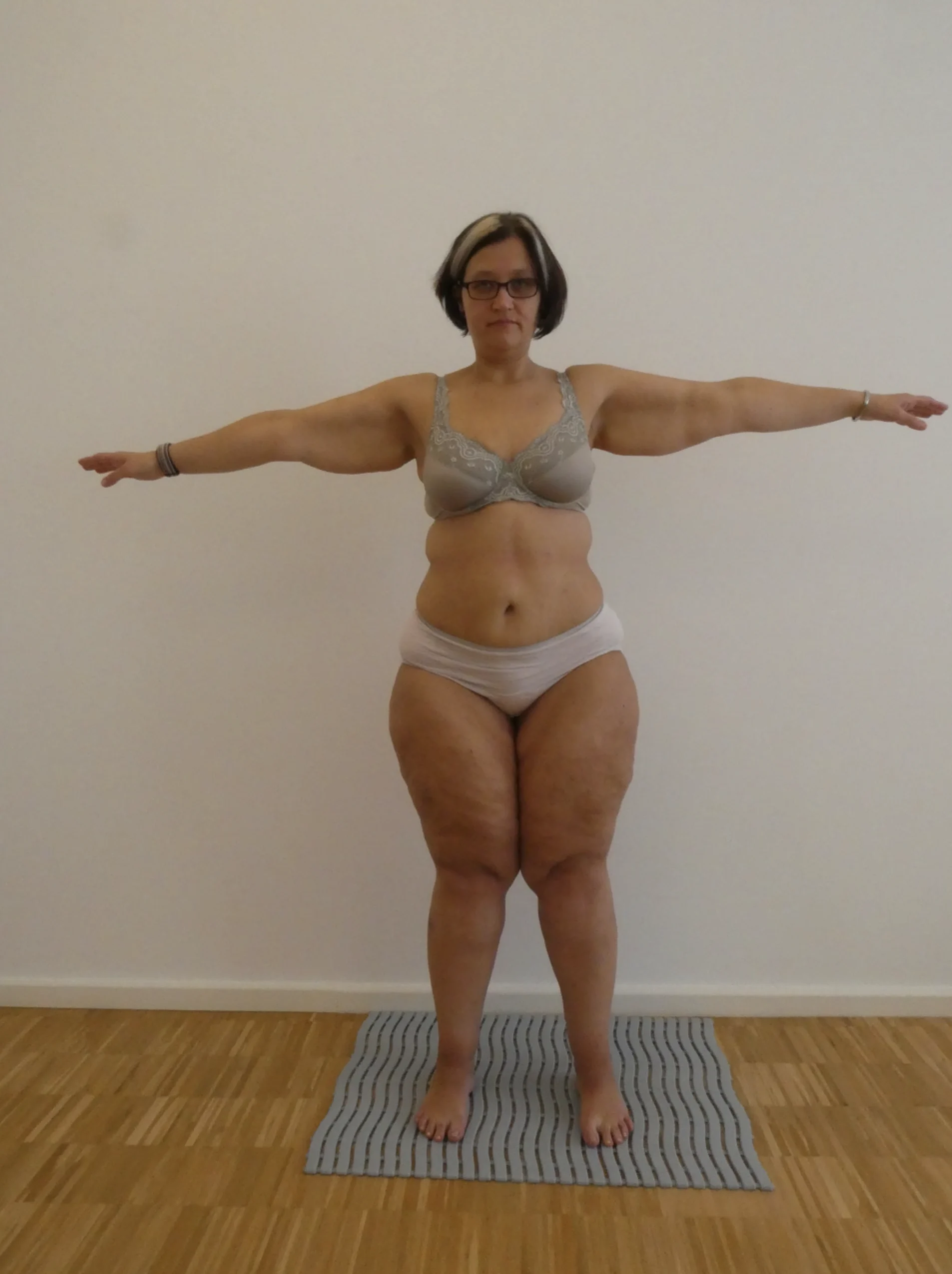 Person in Unterwäsche und Brille steht mit ausgestreckten Armen auf einer Matte in einem gut beleuchteten Raum.