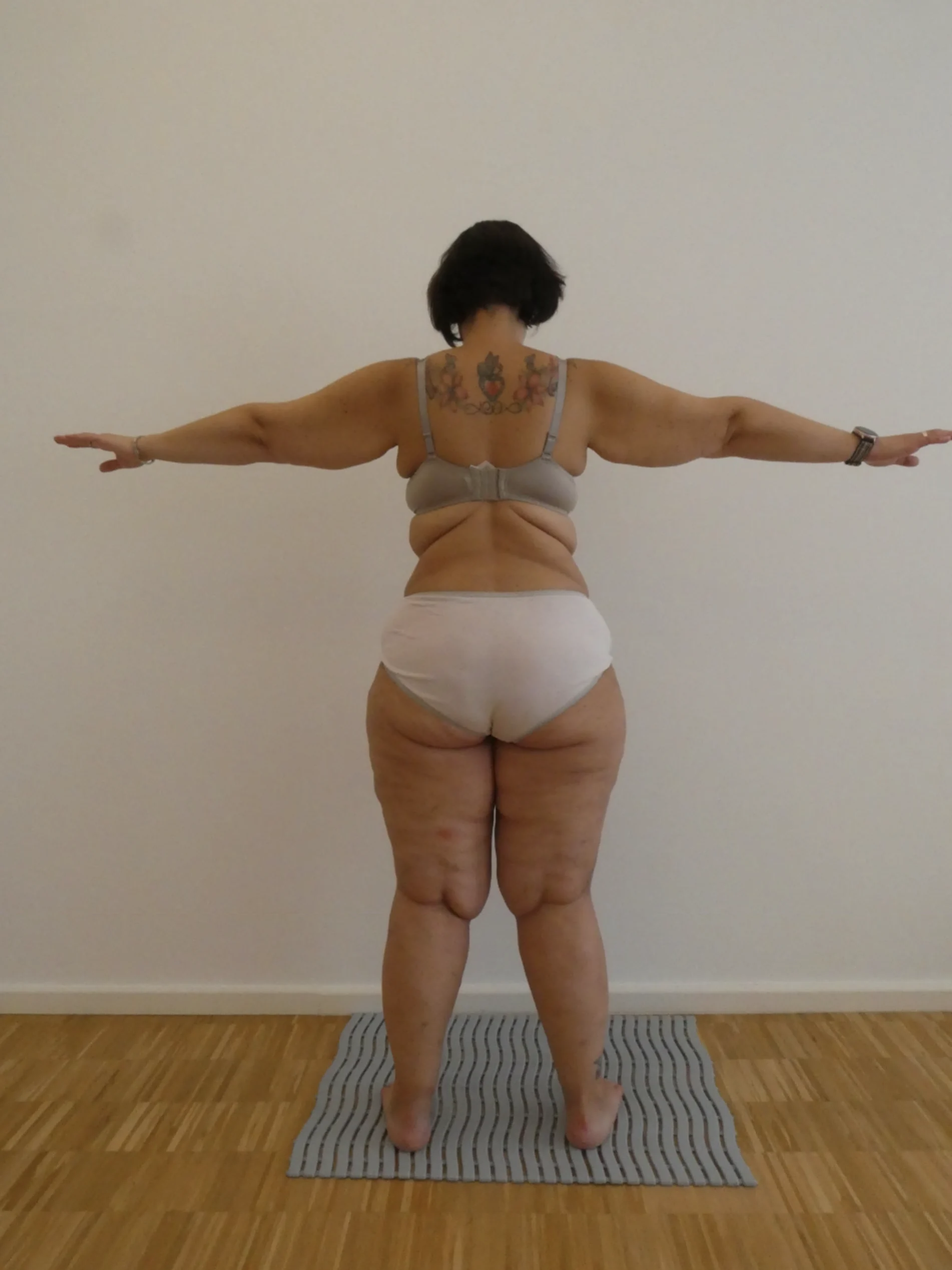 Person mit kurzen Haaren, ausgestreckten Armen, auf gestreifter Matte. Trägt helle Unterwäsche, buntes Rückentattoo; Holzboden.