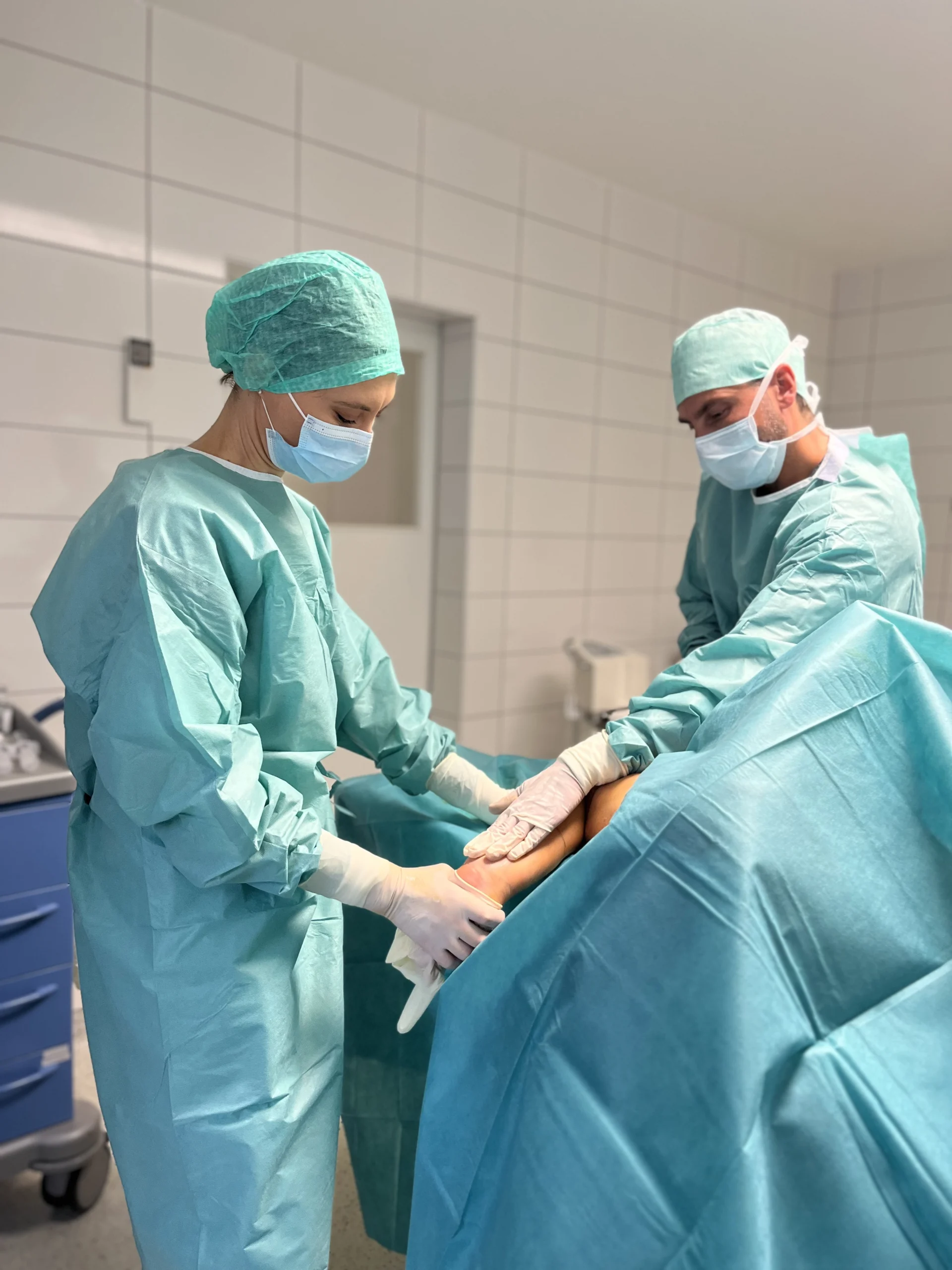 Zwei Chirurgen bereiten im Operationssaal, umgeben von blauen Tüchern in einer sterilen Umgebung, das Bein eines Patienten für die Operation vor.