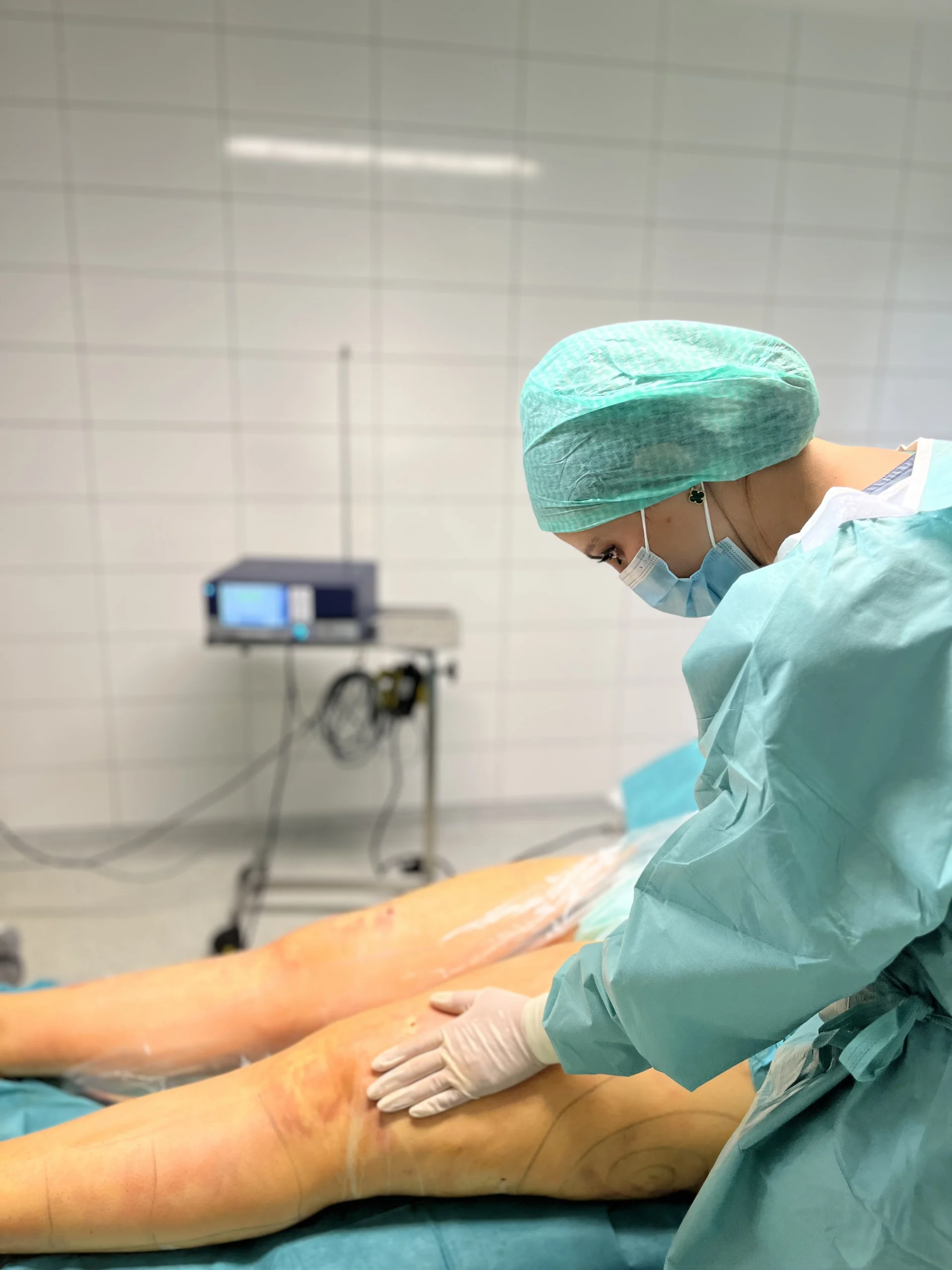 Mediziner in OP-Kleidung untersuchte Patientenbeine auf OP-Tisch; weiß gekachelter Raum mit medizinischer Ausrüstung im Hintergrund.