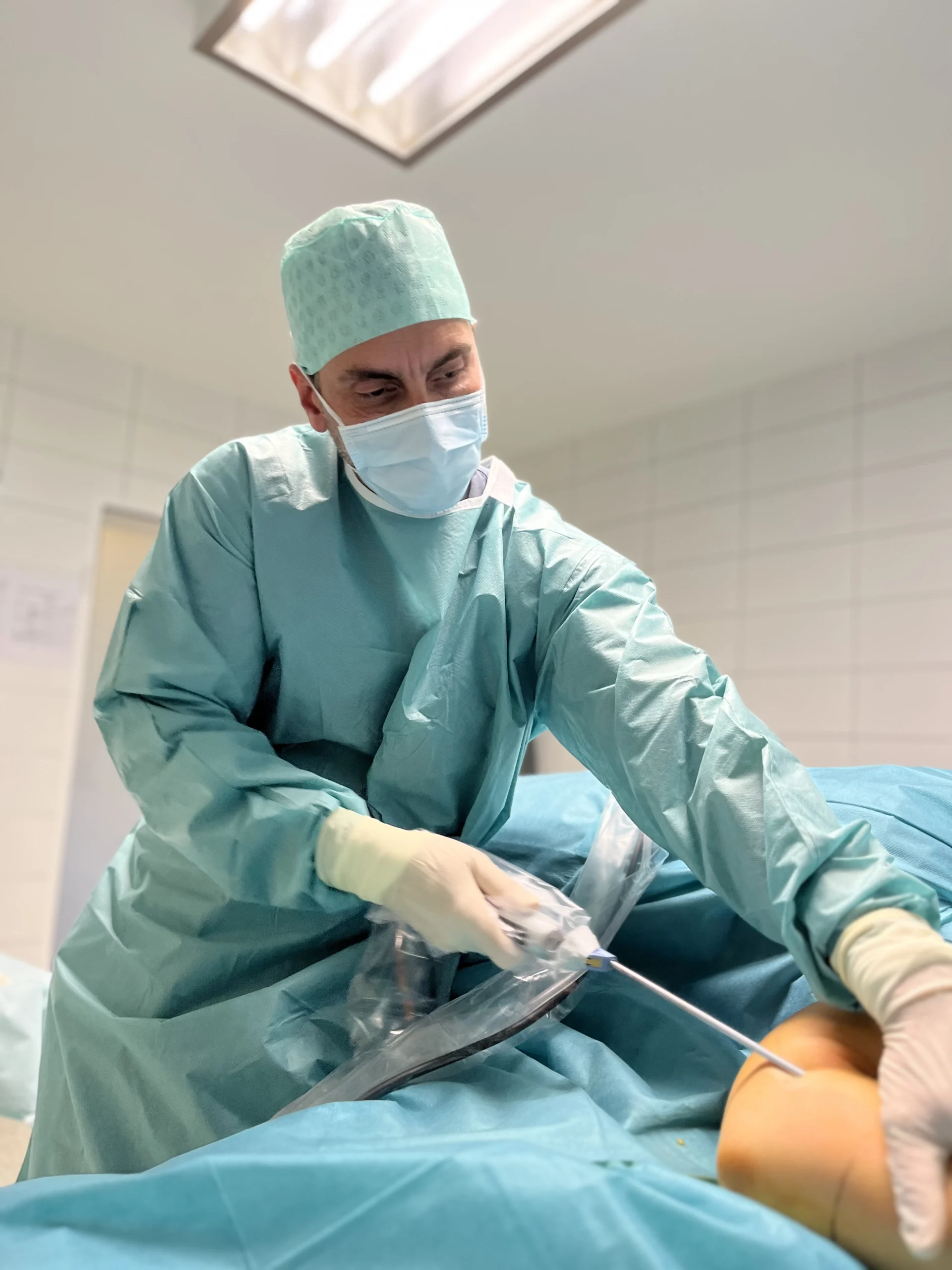 Ein Chirurg im grünen OP-Kittel operiert einen Patienten unter einem blauen Tuch in einem sterilen Raum mit hellem Licht und gefliesten Wänden.