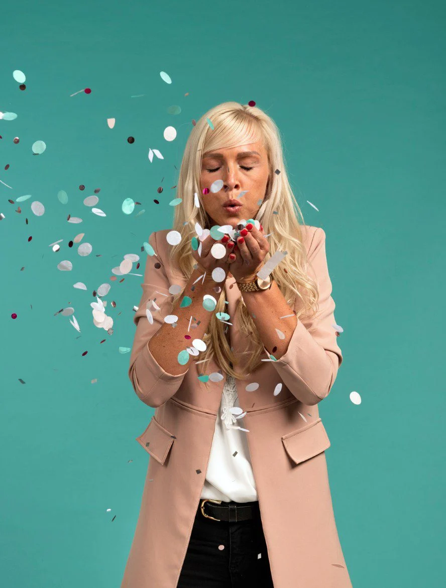 Blonde Frau in beigem Blazer bläst buntes Konfetti vor tealfarbenem Hintergrund, wirkt fröhlich und verspielt.