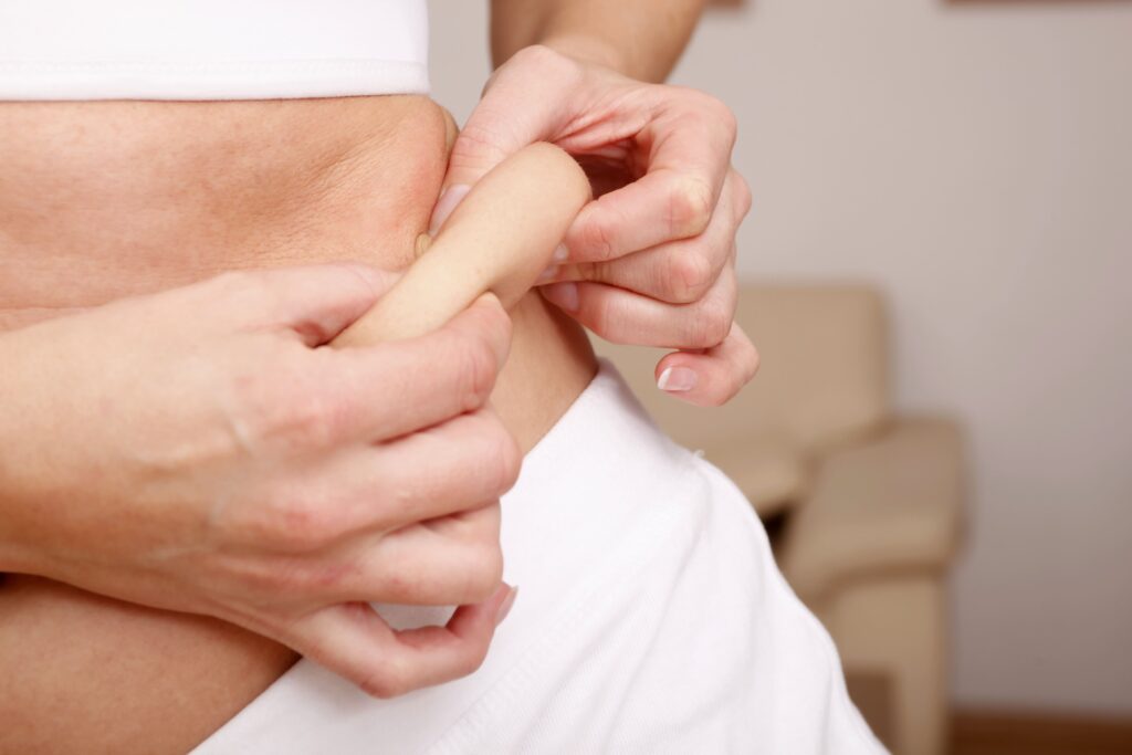 Person in weißer Kleidung kneift Haut am Bauch, denkt an Hüft-Liposuktion, steht drinnen vor neutralen Möbeln.