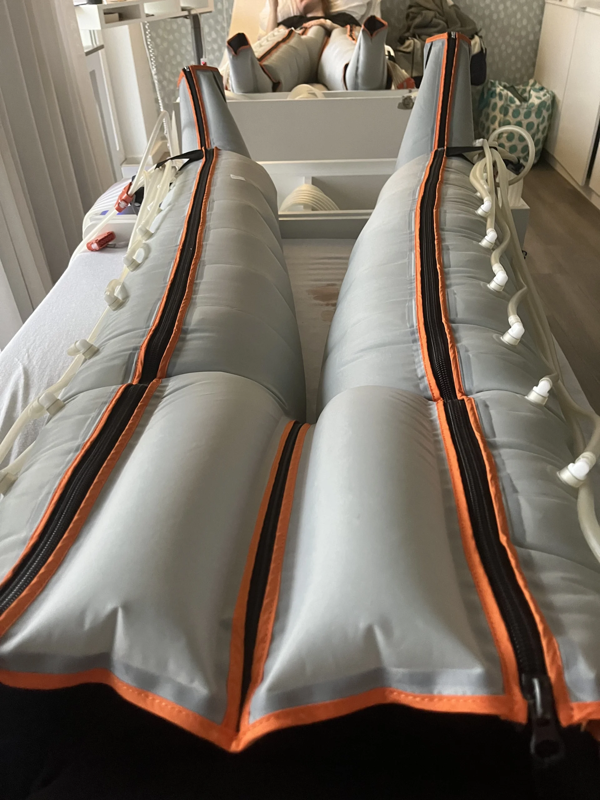 Beine einer Person in orangefarbenen, aufblasbaren Kompressionshüllen mit Reißverschluss von Laura Schabacker, auf einem Bett in einem hellen Raum liegend.