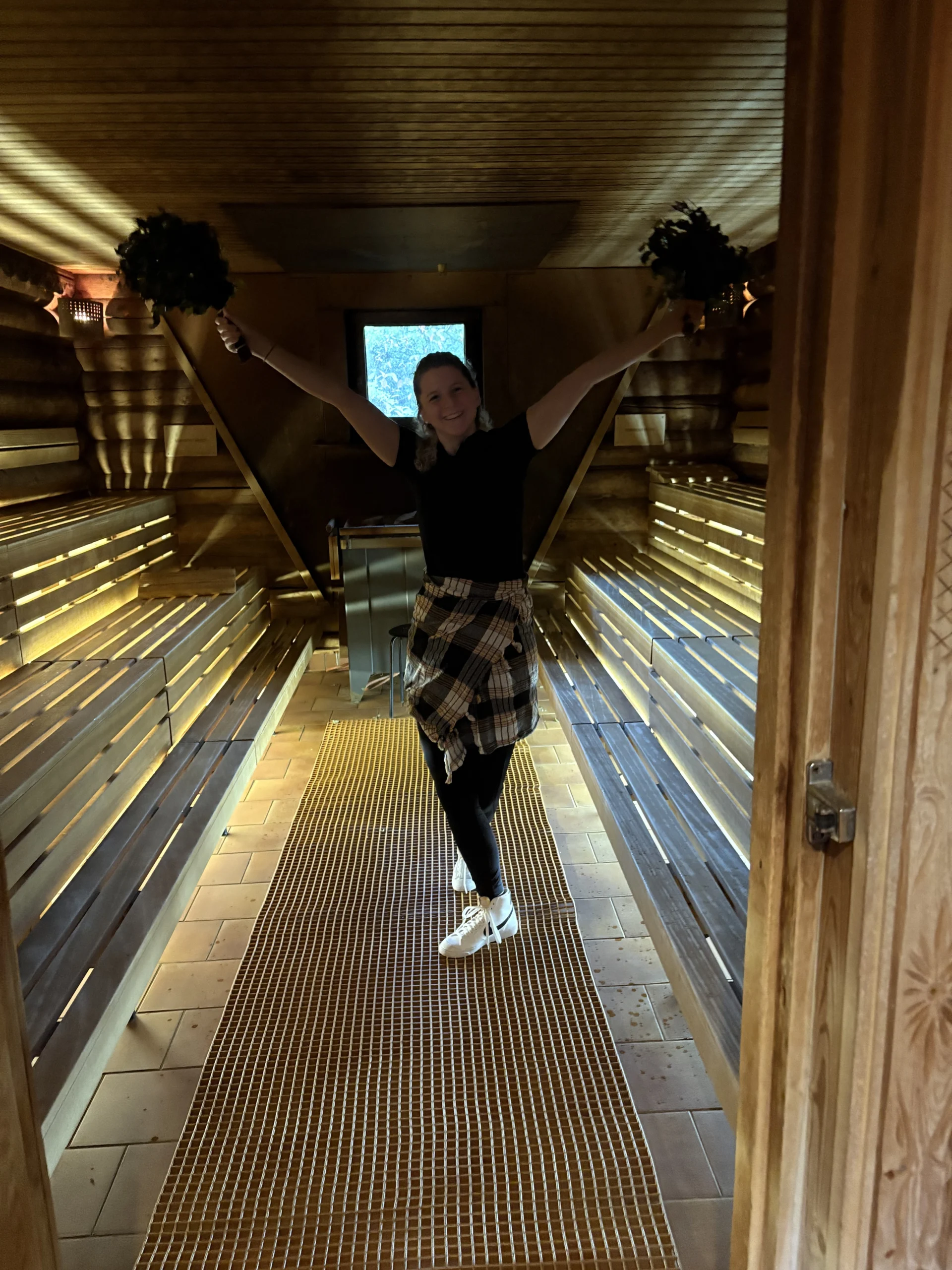 Laura Schabacker lächelt in einer hölzernen Sauna, die Arme mit den belaubten Ästen nach oben gestreckt, lässige Kleidung und warmes Licht umgeben sie.