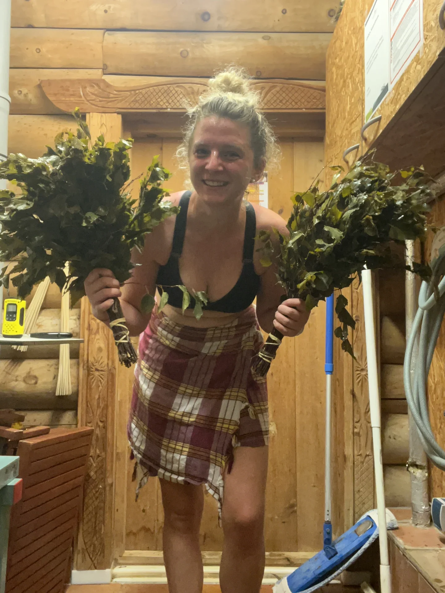 Person mit Lipödem-Erfahrung lächelt drinnen, trägt karierte Decke, schwarzen Top, hält Zweige. Holzige Umgebung.