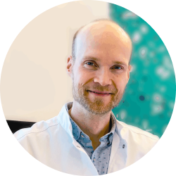 Lächelnder Mann mit Glatze, heller Haut, gestutztem Bart, weißem Mantel, blauem Hemd; blaugrüner und weißer unscharfer Hintergrund.