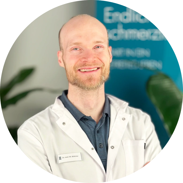 Ein lächelnder, bärtiger Mann in einem weißen Laborkittel mit CG LYMPHA-Namensschild steht vor einer unscharfen blauen Beschilderung und grünen Pflanzen.